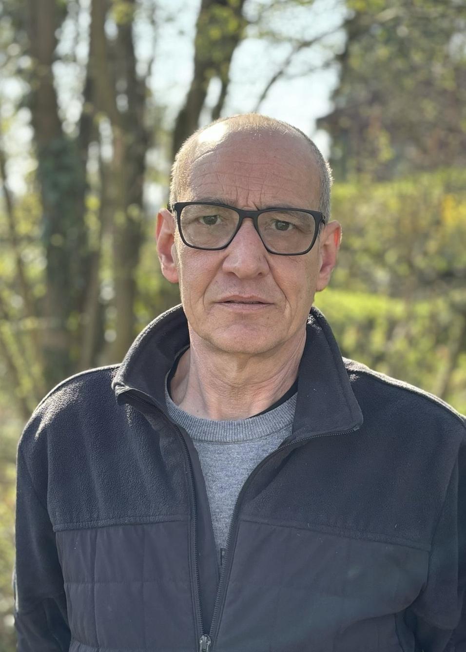 Mann mit Brille in einem schwarzen Fleece-Jackett steht vor einer gr&uuml;nen, bewaldeten Kulisse.