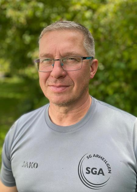 Portrait eines l&auml;chelnden Mannes mit Brille, in grauem Shirt mit Aufdruck, vor einer gr&uuml;nen Pflanzenkulisse.
