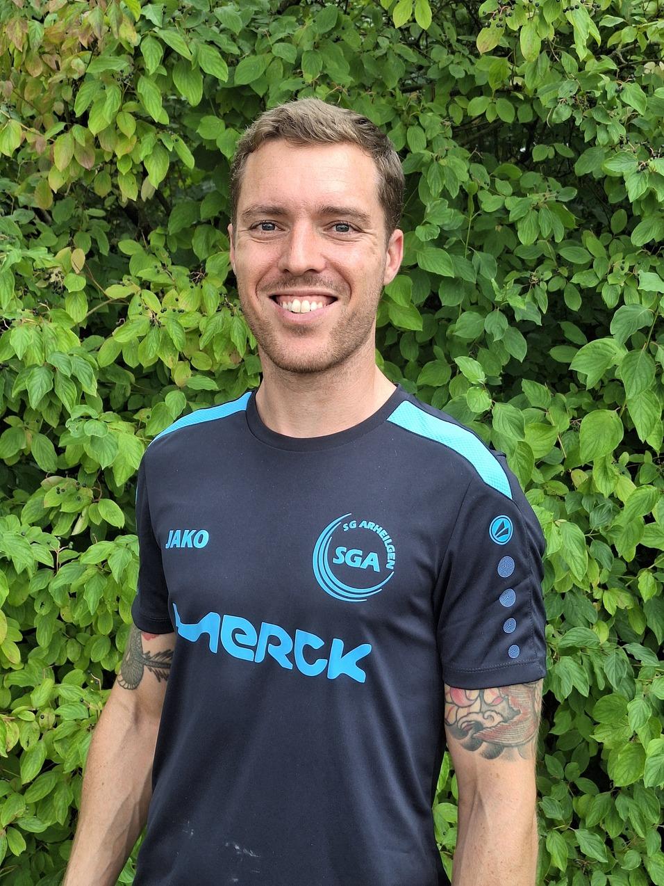 Mann mit kurzen Haaren, Trikot der SGA Arnecken mit blauem Logo, steht vor gr&uuml;nem Hintergrund aus Pflanzen.
