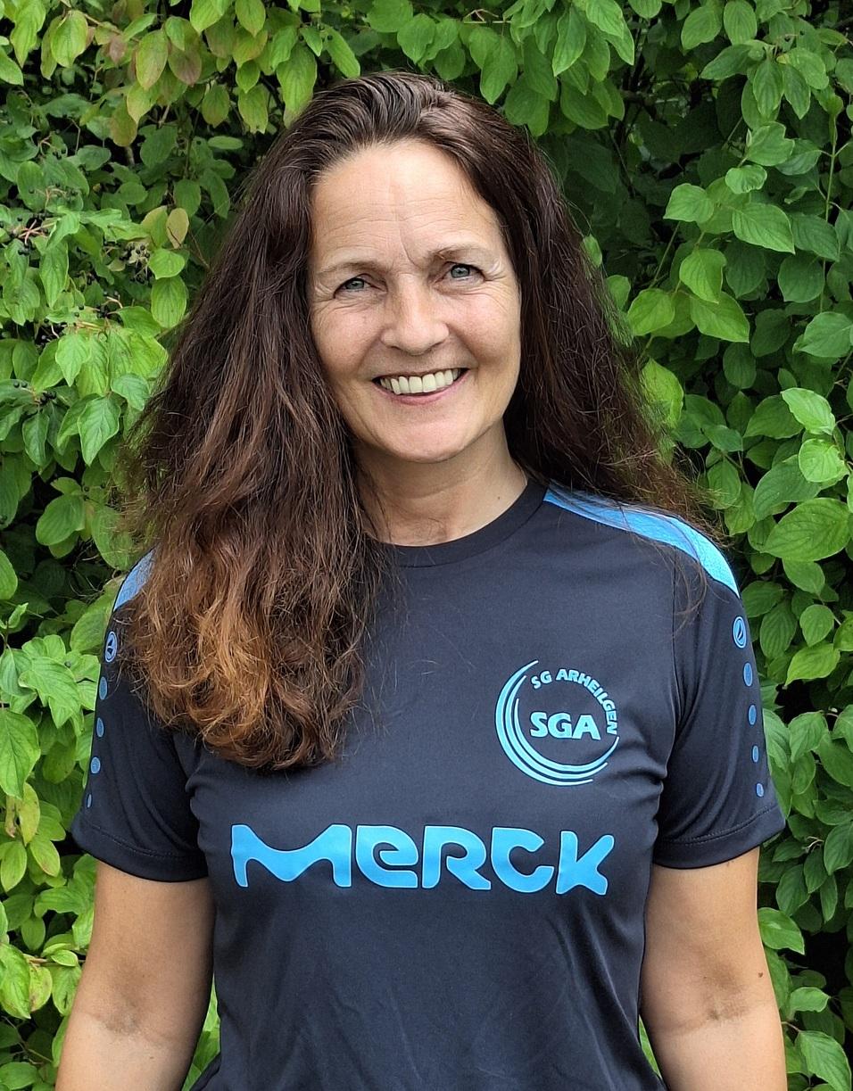 Frau mit langen Haaren tr&auml;gt ein schwarzes Vereins-T-Shirt mit blauer Schrift vor gr&uuml;nem Hintergrund.