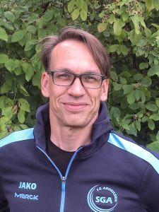 Portr&auml;t eines l&auml;chelnden Mannes mit Brille, der einen Sportjacke tr&auml;gt, vor einem gr&uuml;nen Hintergrund aus Bl&auml;ttern.