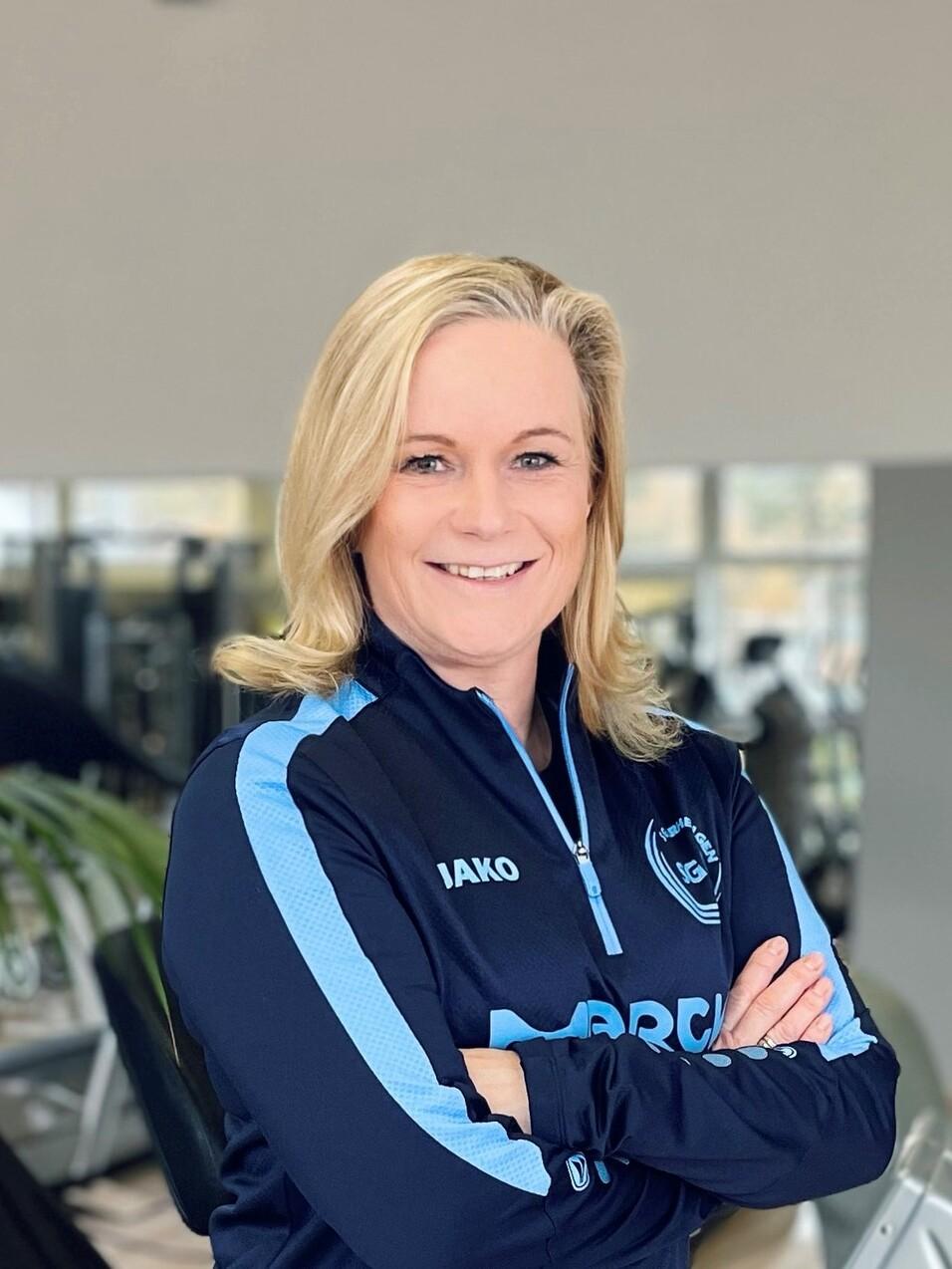 Frau mit blonden Haaren, in Sportbekleidung, l&auml;chelt mit verschr&auml;nkten Armen in Fitnessstudio-Hintergrund.