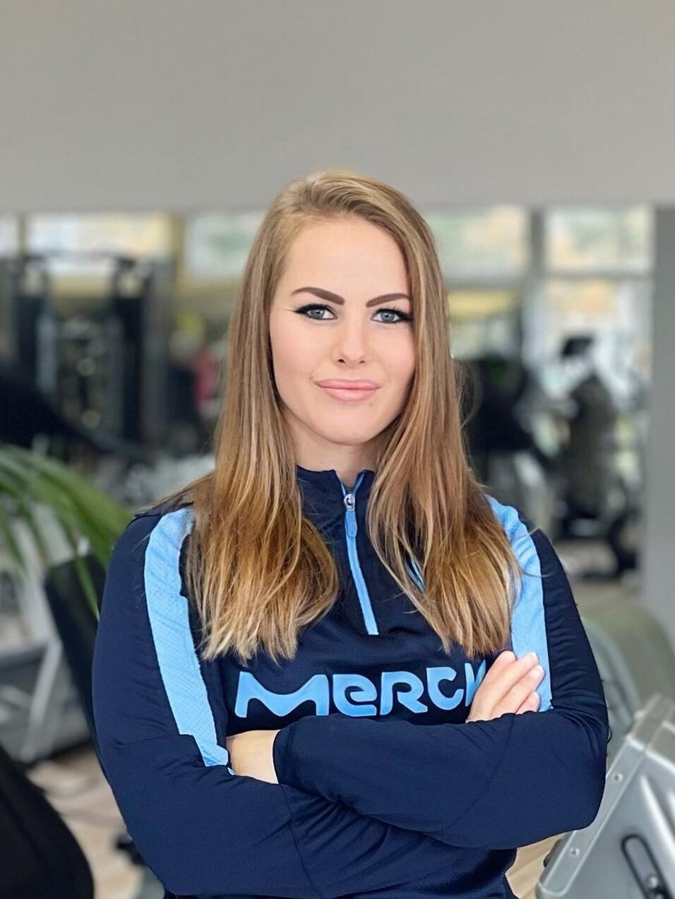 Frau mit langen, braunen Haaren in sportlicher Kleidung steht in einem Fitnessstudio mit verschr&auml;nkten Armen.