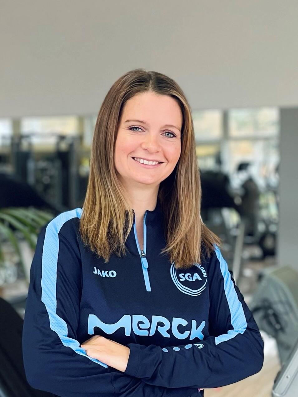 Portrait einer l&auml;chelnden Frau in Sportbekleidung mit dem Logo &bdquo;SGA Merck&ldquo;, stehend in einem Fitnessstudio.