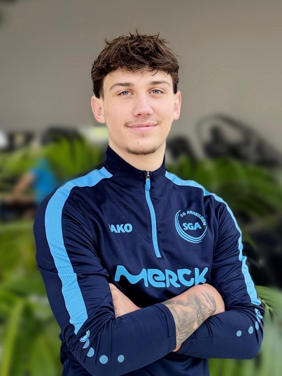 Junger Mann in Sportjacke mit l&auml;chelndem Gesicht, Arme vor der Brust gekreuzt, Hintergrund mit gr&uuml;nen Pflanzen.