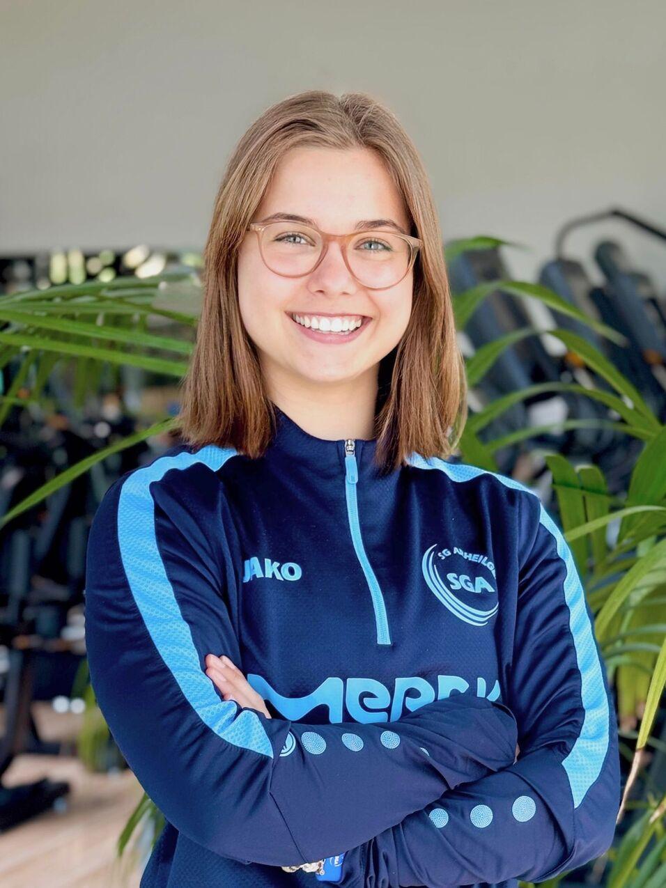 L&auml;chelnde Frau mit Brille in blauer Sportbekleidung, Arme vor der Brust verschr&auml;nkt, Hintergrund mit Pflanzen und Fitnessger&auml;ten.
