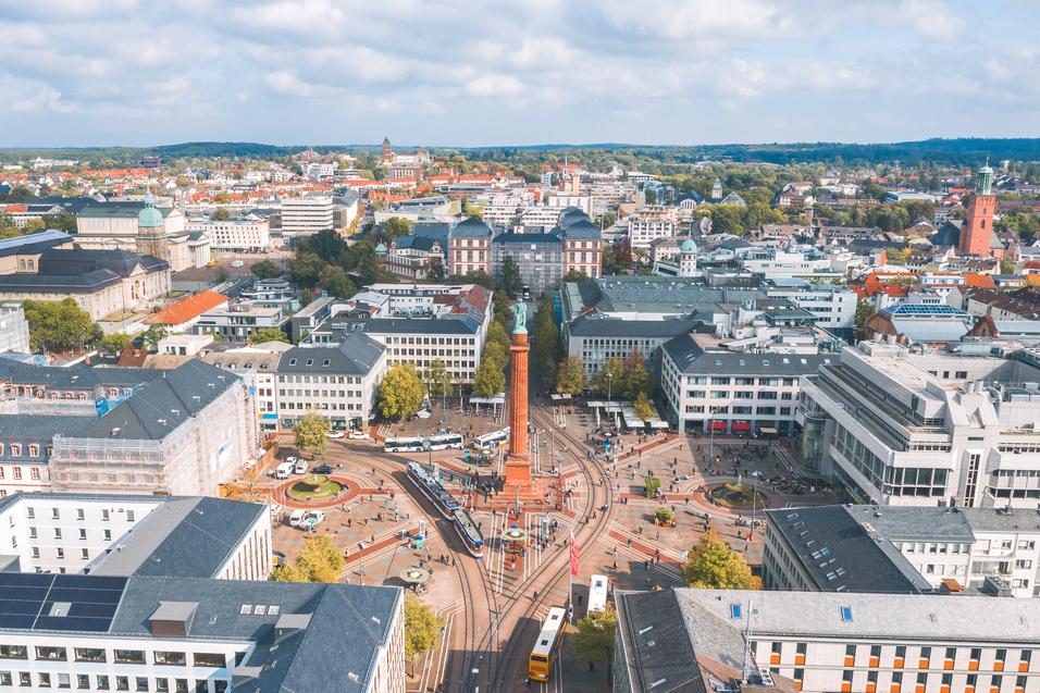 Blick auf eine belebte Innenstadt mit Platz, Stra&szlig;enbahnen und modernen Geb&auml;uden unter einem bew&ouml;lkten Himmel.