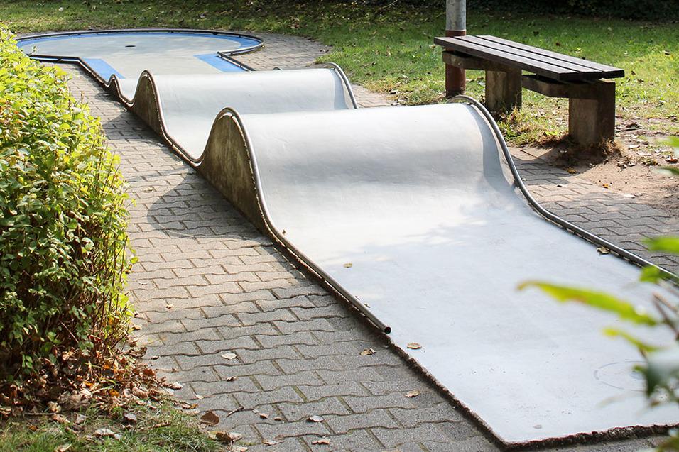 Landschaft mit Wellenbahn f&uuml;r Kinder auf einem gepflasterten Weg, neben einer Bank und gr&uuml;ner Bepflanzung.