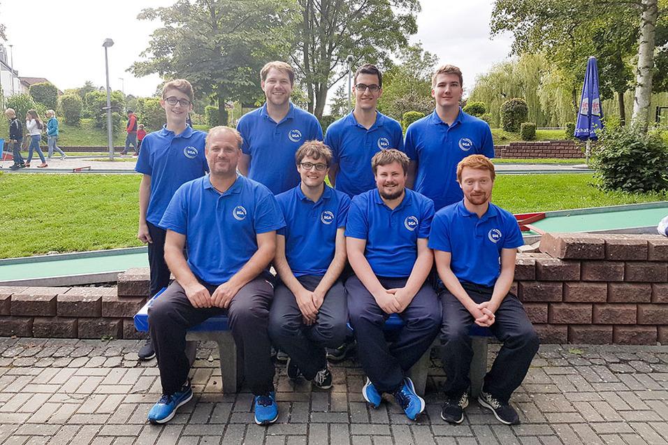 Gruppenbild von acht Personen in blauen T-Shirts, sitzend und stehend auf einer Bank im Freien, mit gr&uuml;nem Hintergrund.