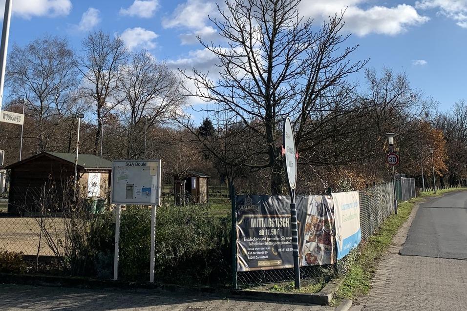 Eingang zu einem Park mit B&auml;umen, Schildern und einem Zaun, im Hintergrund eine kleine Holzh&uuml;tte.