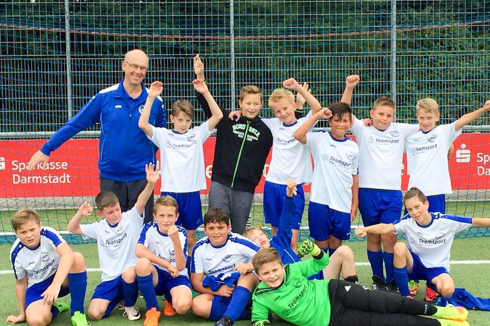 Gruppe von 12 Kindern in Fußballtrikots, posierend mit Trainer auf einem Sportplatz im Freien.