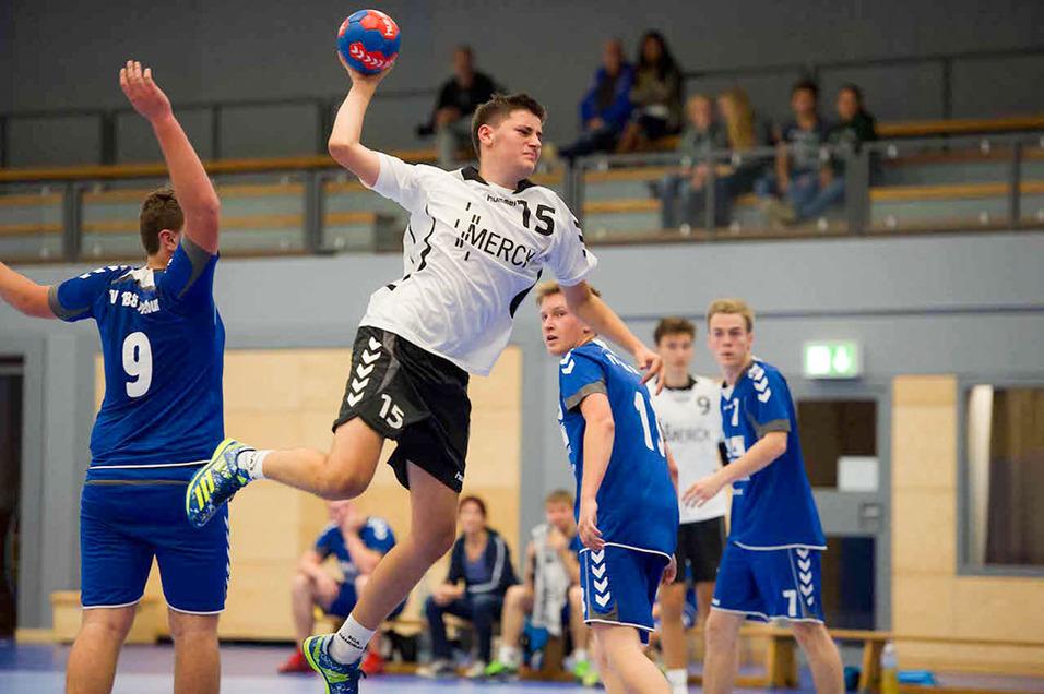 Ein Handballspieler in wei&szlig;em Trikot springt, um den Ball auf ein Tor zu werfen, w&auml;hrend Gegner in blauen Trikots reagieren.