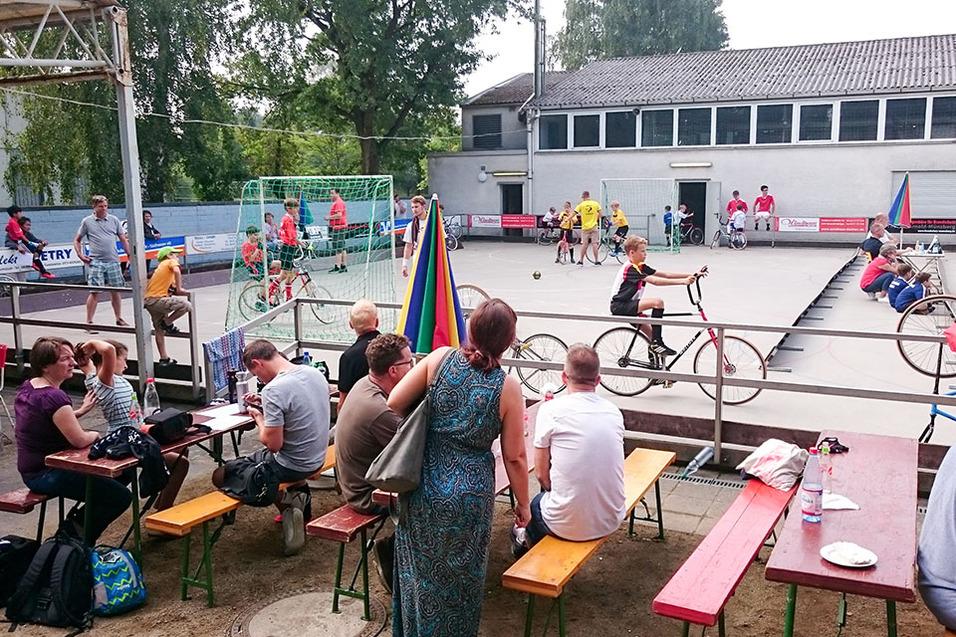 Menschen beobachten ein Spiel auf einem Sportplatz, während Kinder mit Fahrrädern und Bällen aktiv sind.