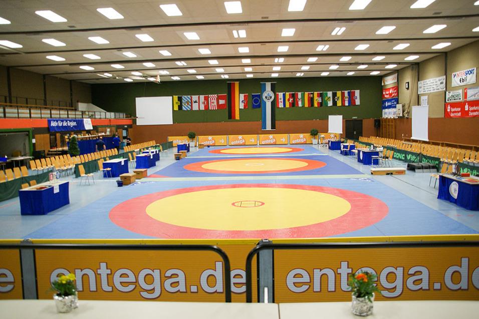 Blick auf eine Wettkampfhalle mit Matten f&uuml;r Ringen, Sitzpl&auml;tzen und Nationalflaggen an der Wand.