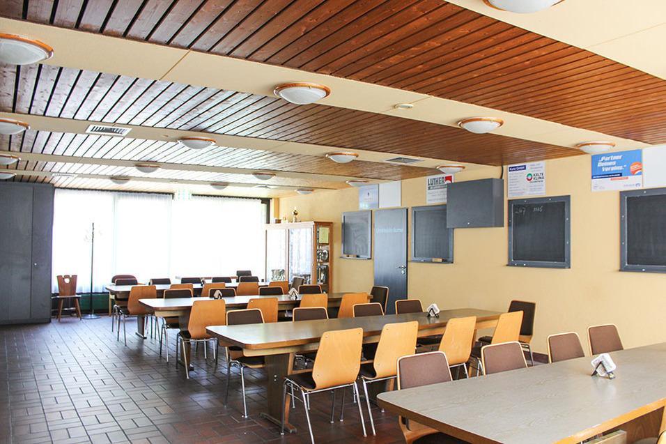 Hell erleuchteter Raum mit Holzdecke, Tischen und St&uuml;hlen, sowie einer gro&szlig;en Fensterfront und mehreren Wandtafeln.