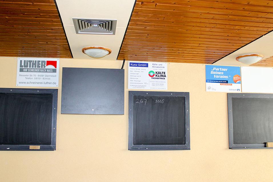 Wand mit Werbeschildern und zwei leeren Tafeln in einem Raum, Holzdecke und Deckenleuchte sichtbar.