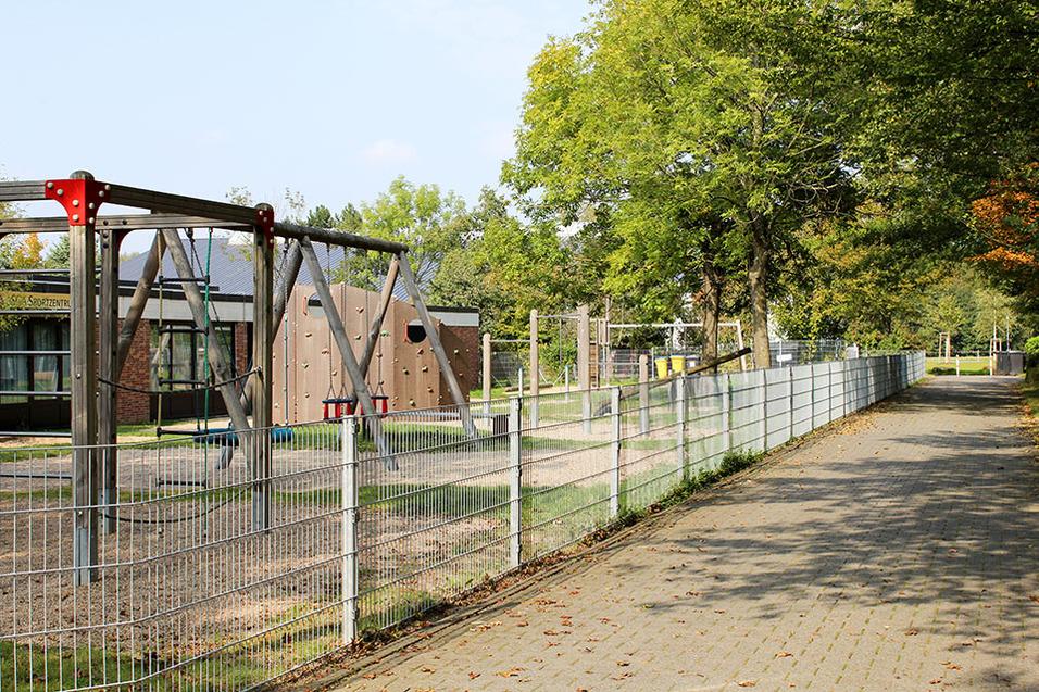 Spielplatz mit Klettergerüst, Schaukeln und einem befestigten Weg, umgeben von Bäumen und einem Zaun.