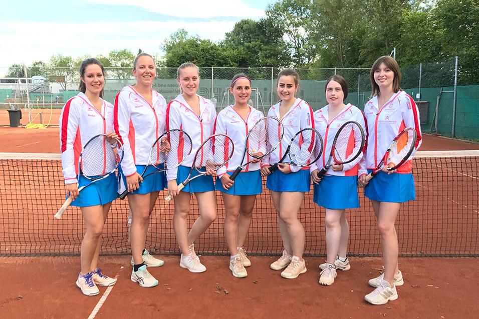 Sieben Tennis-Spielerinnen in einheitlicher Kleidung stehen am Netz eines Tennisplatzes, jede h&auml;lt einen Schl&auml;ger.