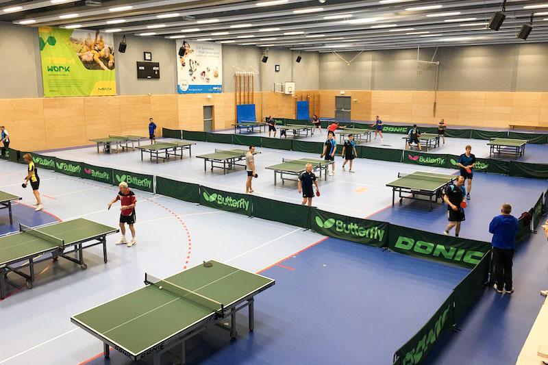 Mehrere Tischtennisspieler trainieren in einer Sporthalle mit mehreren Tischtennisplatten und gr&uuml;nen Umrandungen.
