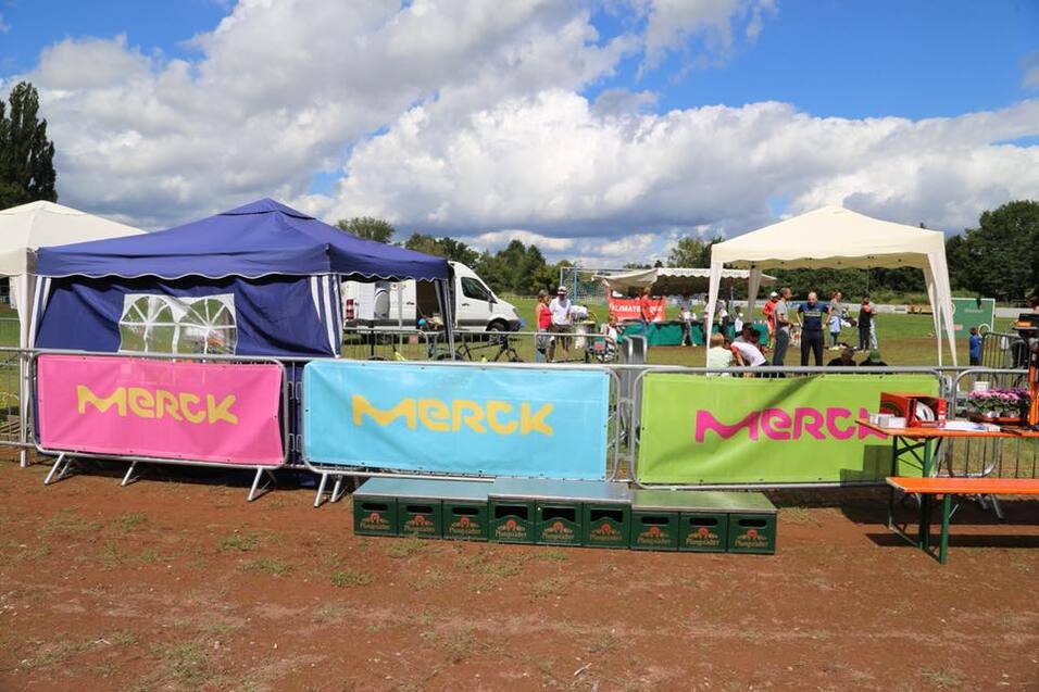 Zwei Zelte mit Merck-Werbung stehen auf einem Sportfeld, umgeben von mehreren Personen und Veranstaltungen.