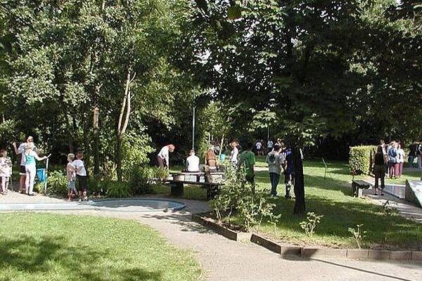 Menschen verschiedenen Alters verbringen Zeit in einem grünen Park mit Bäumen und Wegen, einige spielen oder unterhalten sich.