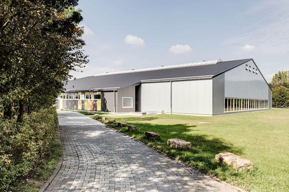 Moderne Sporthalle mit grauem Dach und großen Fenstern, umgeben von Rasen und Bäumen, Weg führt zum Eingang.