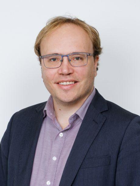 Portr&auml;t eines Mannes mit Brille, kurzem Haar und einem l&auml;chelnden Gesicht, gekleidet in hellblauem Hemd und dunklem Blazer.