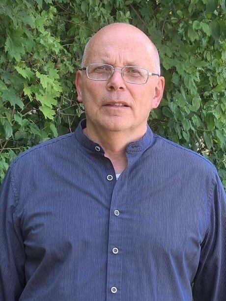 Portr&auml;t eines Mannes mit Glatze und Brille, der ein blaues Hemd tr&auml;gt, vor einem Hintergrund aus gr&uuml;nen Bl&auml;ttern.