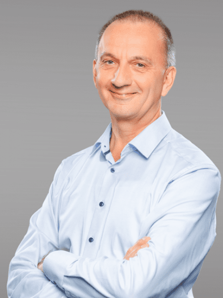 Mann mit kurzem, grauem Haar, tr&auml;gt ein hellblaues Hemd, l&auml;chelt und steht mit verschr&auml;nkten Armen vor einem grauen Hintergrund.