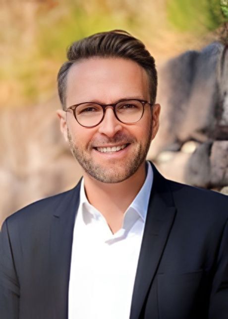 Portr&auml;t eines Mannes mit Brille in einem schwarzen Anzug, l&auml;chelnd vor einem unscharfen Hintergrund.