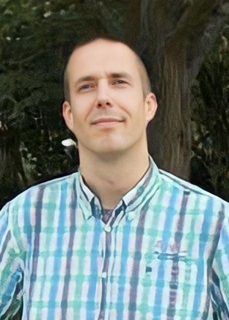 Ein Mann in einem karierten Hemd steht l&auml;chelnd vor einem verschwommenen, gr&uuml;nen Hintergrund.