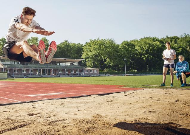 Weitspringer in der Luft beim Absprung über eine Sandgrube, daneben zwei Beobachterinnen.