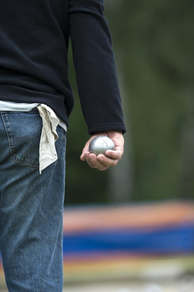 Mann in schwarzem Sweatshirt hält eine Metallkugel in der rechten Hand, während er auf einen Spielplatz schaut.
