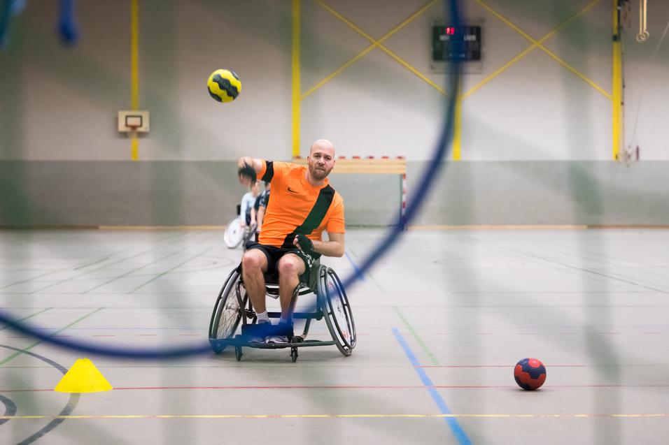 Rollstuhlsportler wirft einen gelben Handball in einer Sporthalle, mit einem blauen Ball und Markierungscones im Hintergrund.