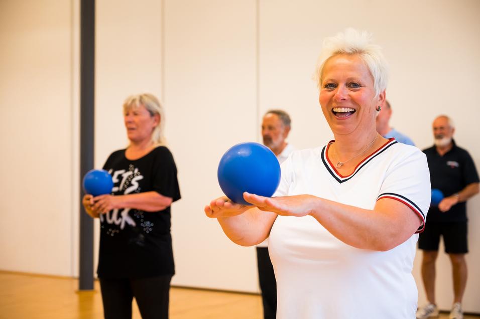 Gruppe älterer Menschen bei einer Übung mit blauen Bällen in einem hellen Raum. Fokus auf lächelnde Frau vorne.