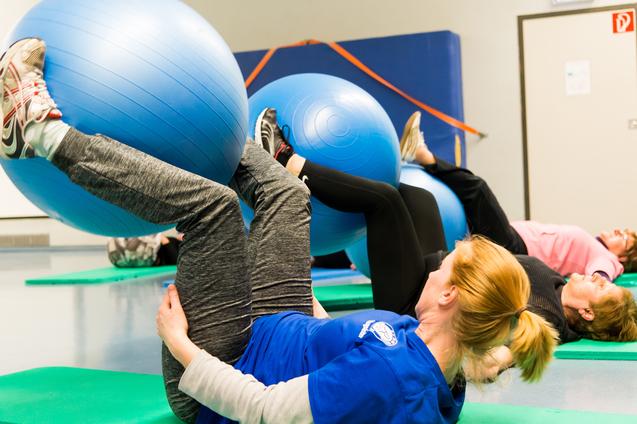 Drei Frauen liegen auf Matten und trainieren mit blauen Gymnastikbällen in einer Sporthalle.
