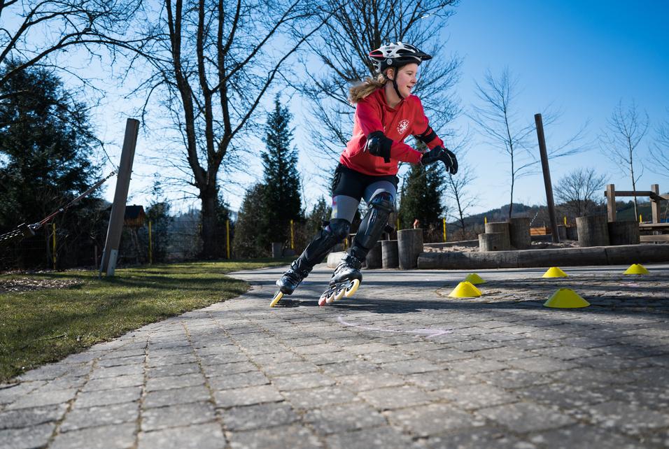 Mädchen in Schutzkleidung fährt auf Inline-Skates und weicht gelben Hütchen auf einem Pflasterweg aus.