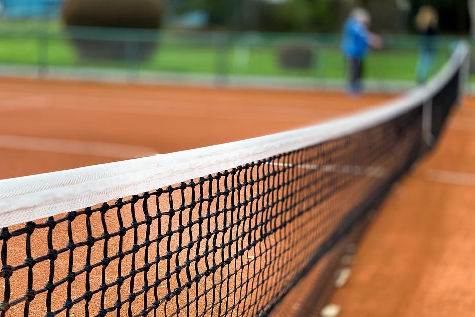 Nahaufnahme eines Tennisnetzes auf einem roten Sandplatz, Hintergrund unscharf mit grünen Bäumen.