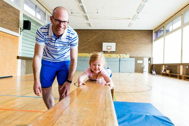 Mann lehnt am Schwebebalken, während ein lächelndes Mädchen darauf liegt, in einer hellen Sporthalle.